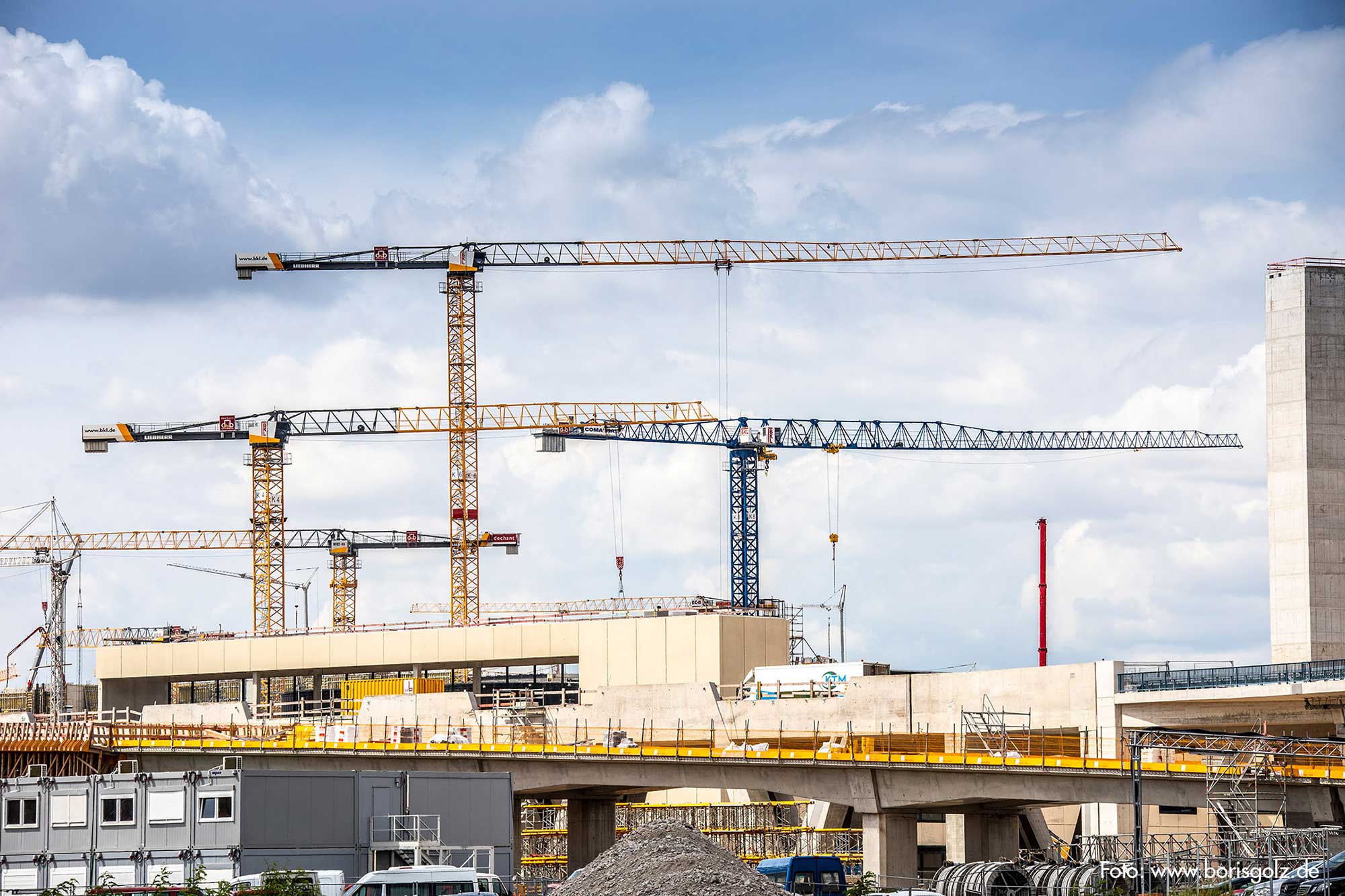 Several BKL construction cranes, including yellow Liebherr cranes, on a large construction site with shell buildings and construction containers, illustrating the construction progress and the comprehensive services of BKL crane rental and logistics.