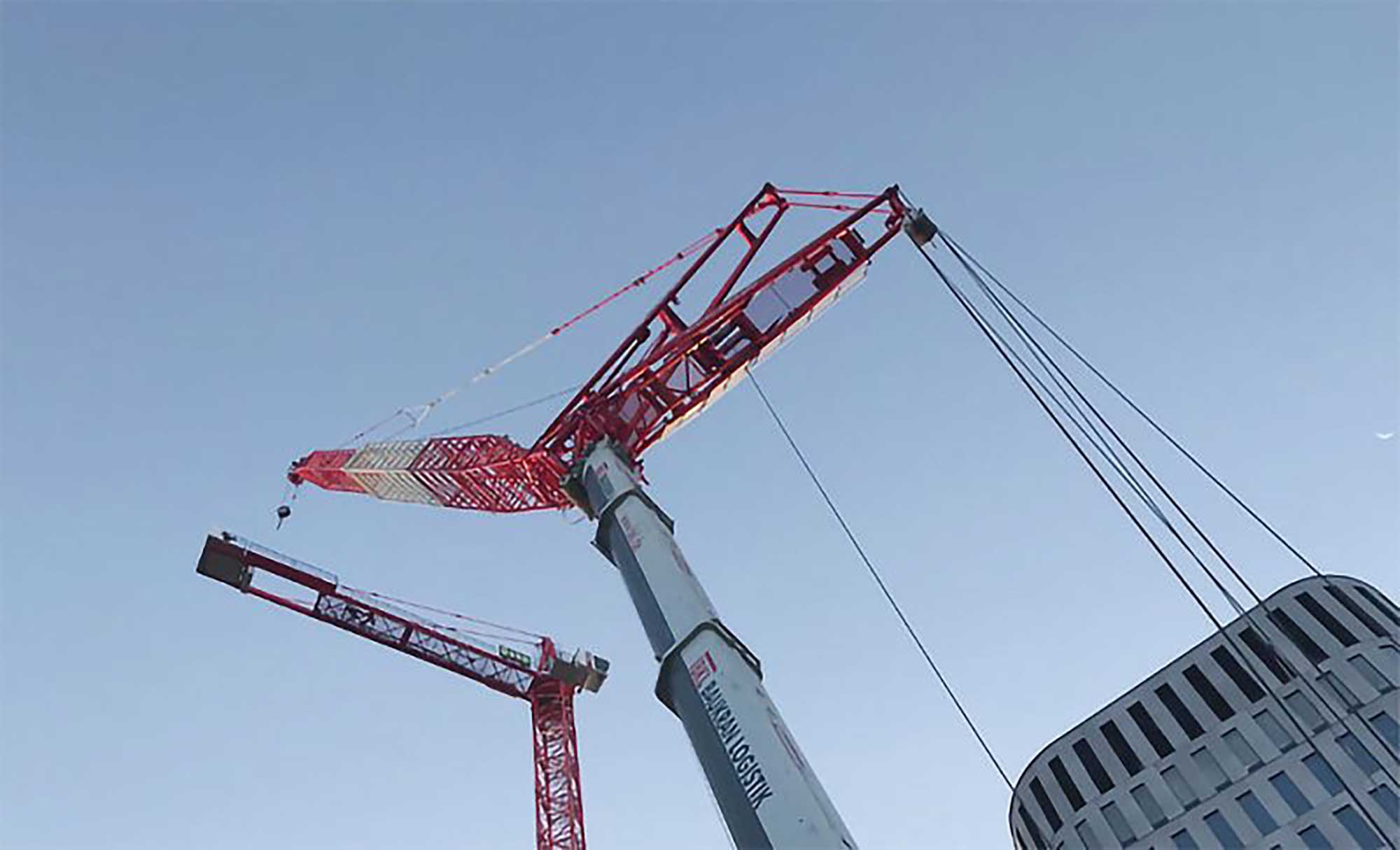 BKL mobile crane during crane assembly on construction site, lifting lattice mast section next to red tower crane and modern building under blue sky.