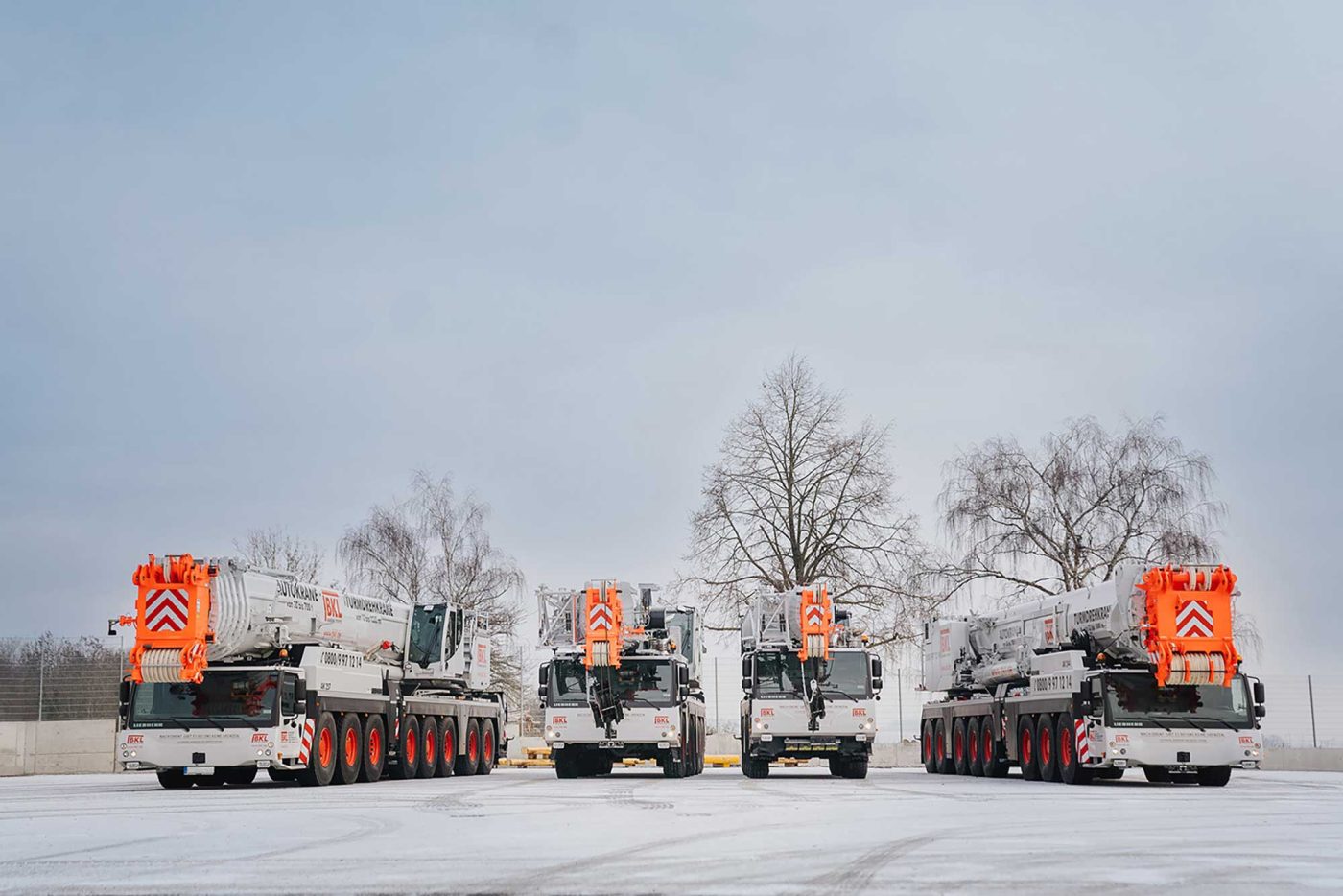 Vier Mobilkrane von BKL Baukran Logistik, weiß mit orangefarbenen Details, auf einem schneebedeckten Betriebsgelände im Winter