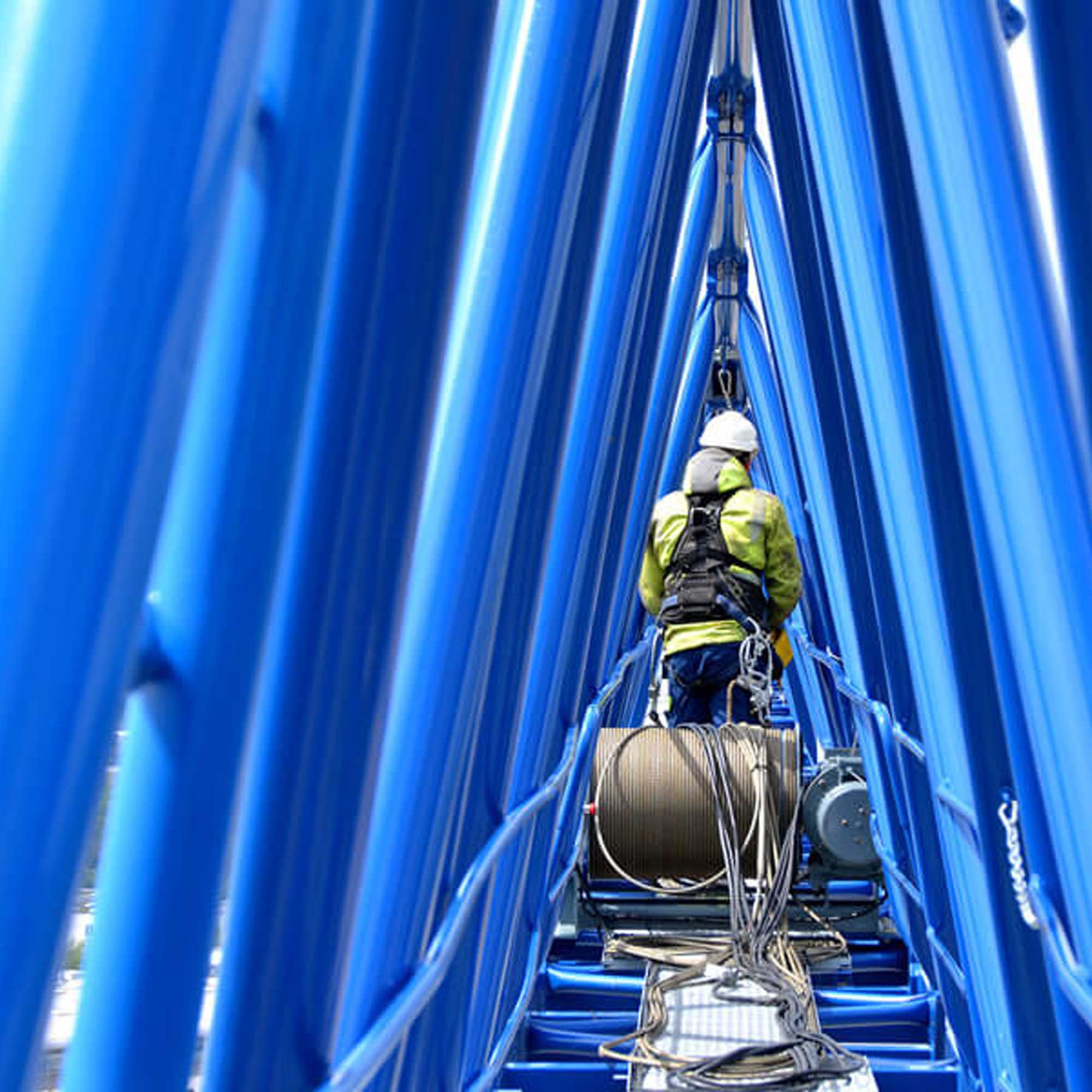 BKL Kranservice-Techniker des 24-Stunden-Notdienst mit Sicherheitsausrüstung bei Wartungs- oder Reparaturarbeiten an der blauen Trägerkonstruktion eines Baukrans, mit sichtbarer Seilrolle.