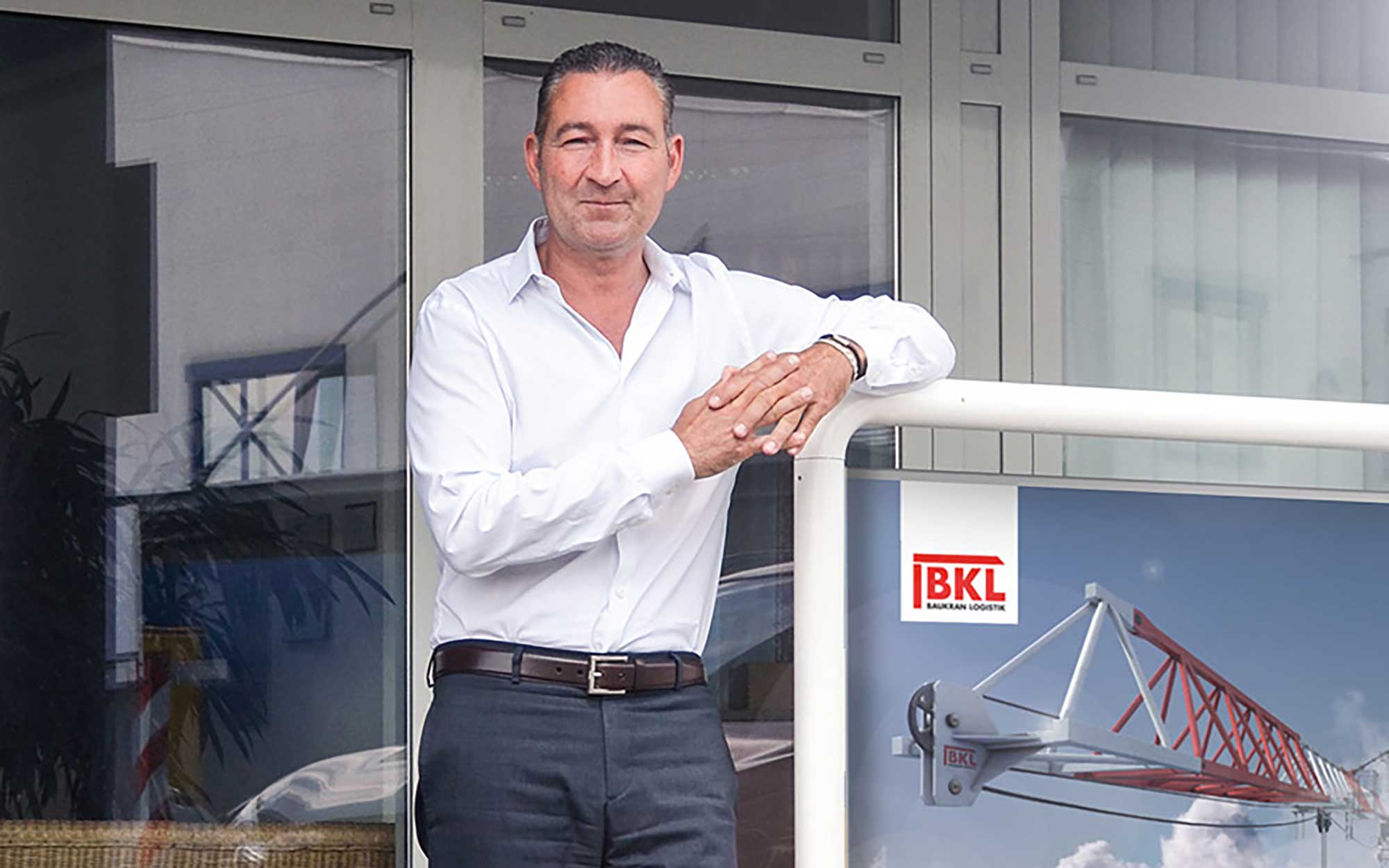 Alexander Volz, Geschäftsführender Gesellschafter, der BKL Baukran Logistik, dem Familienunternehmen für Kranvermietung und Kranlogistik, steht vor dem modernen Firmengebäude. Zu sehen ist auch das BKL Logo und ein Baukran.