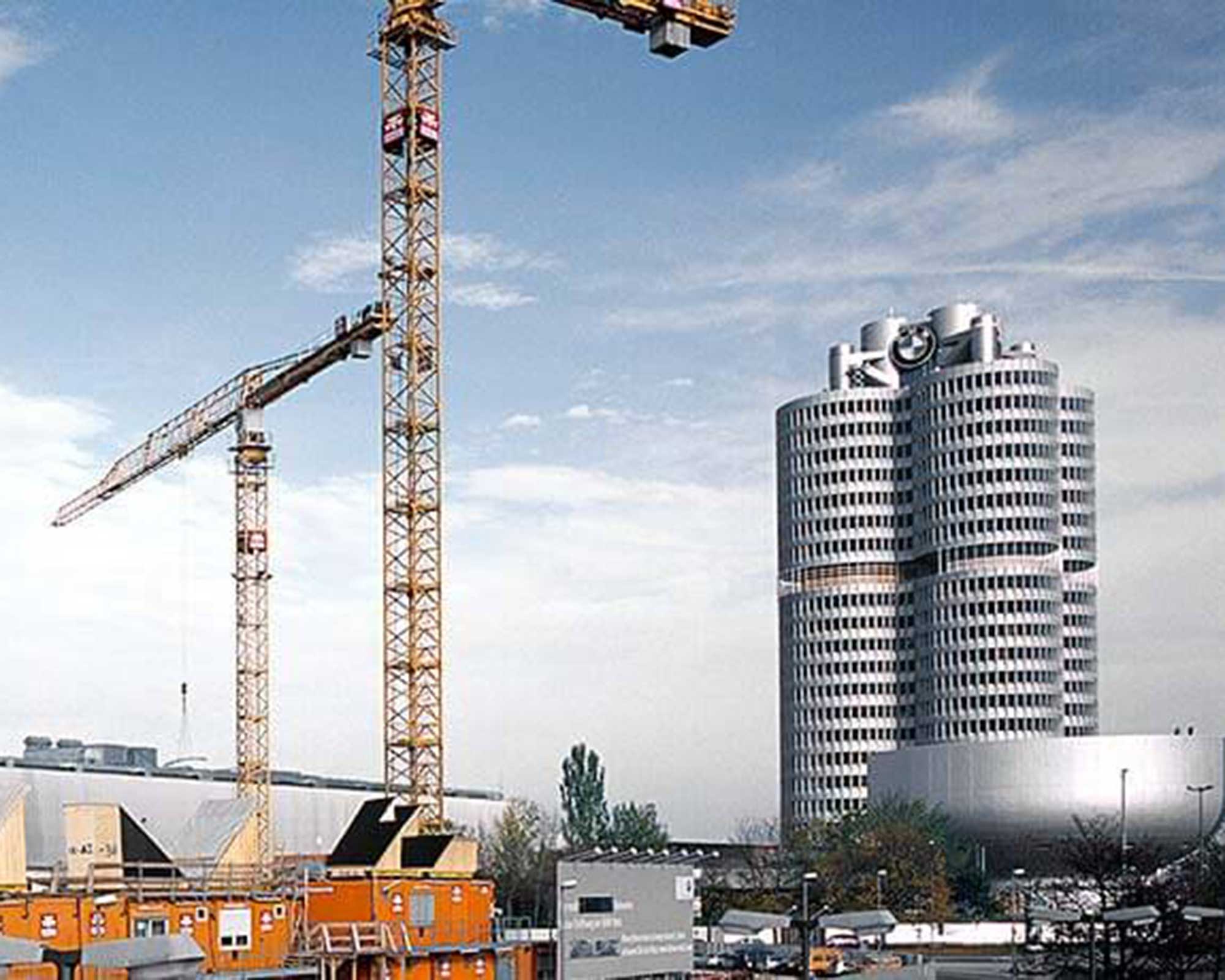Zeigt eine BKL Referenz und den BKL Einsatz für die BMW Welt: Zwei gelbe Liebherr Turmdrehkrane 280 EC-H und EC-B Litronic von BKL auf Stahlgerüsten an der Baugrube der BMW Welt in München, mit dem BMW Vierzylinder-Hochhaus im Hintergrund.