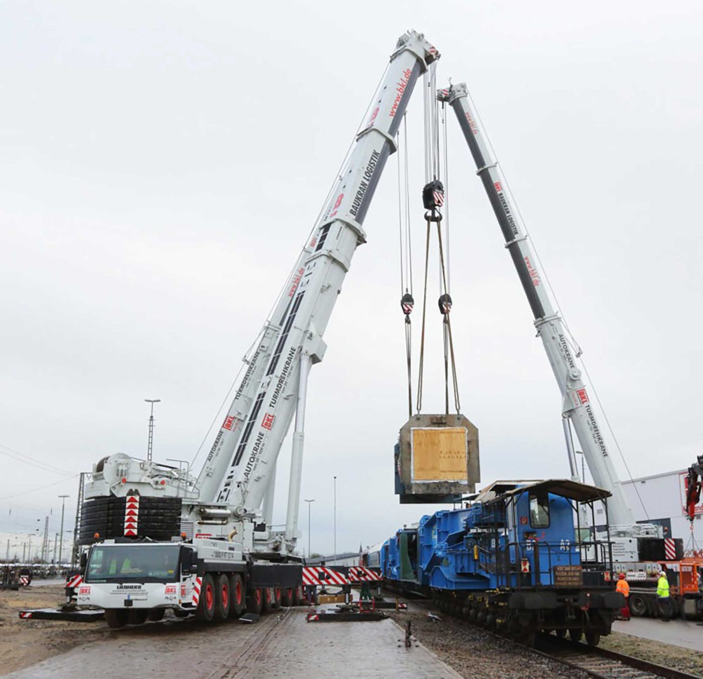 Zwei BKL Mobilkrane im Tandemhub verladen Schwerlast auf blaue Lokomotive für Schienentransport und Speziallogistik