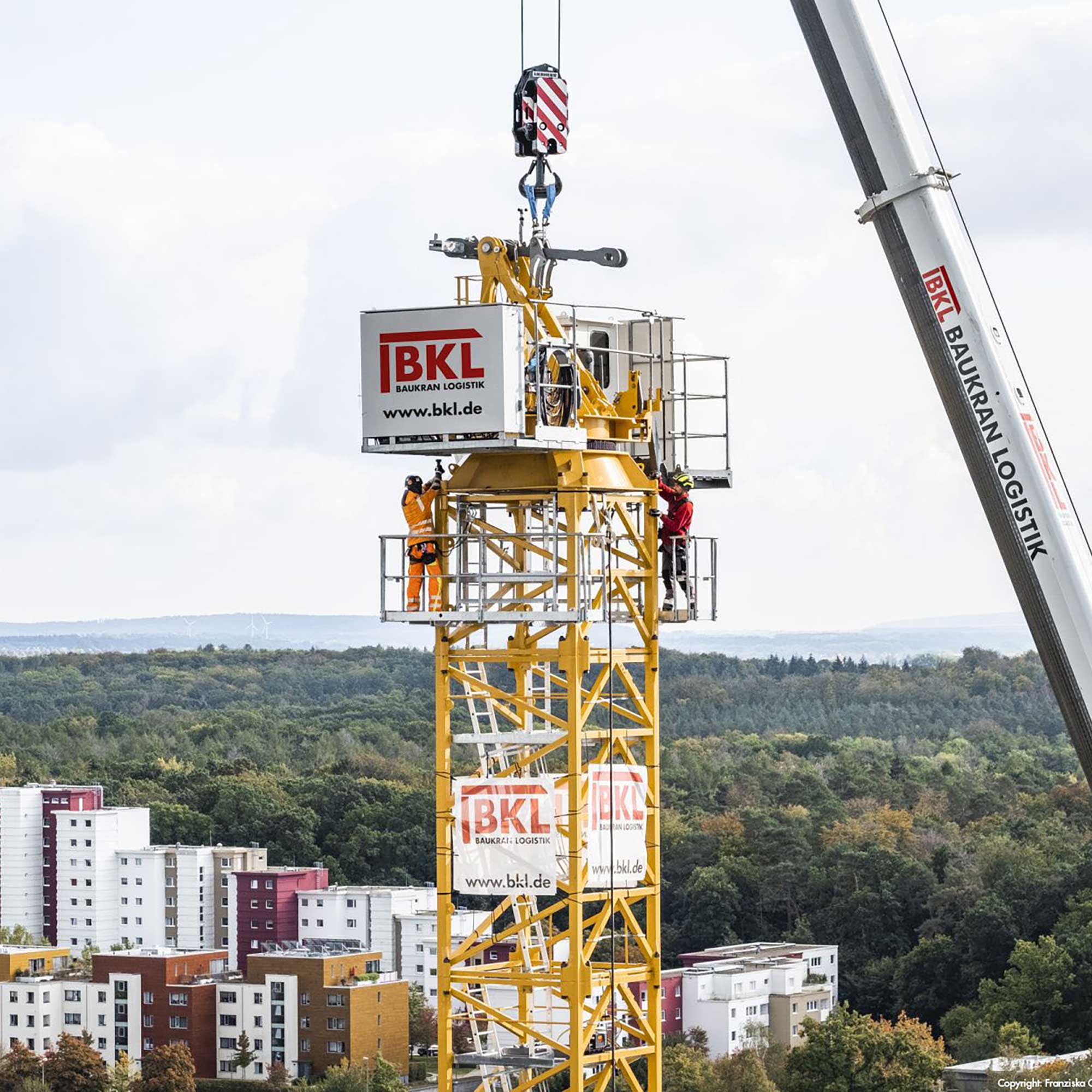 Zwei Techniker in Schutzkleidung montieren einen gelben Turmdrehkran in großer Höhe, unterstützt von einem Mobilkran, über einer Stadt- und Waldlandschaft. Dieses Bild demonstriert den professionellen BKL Kranservice für Kranmontage.