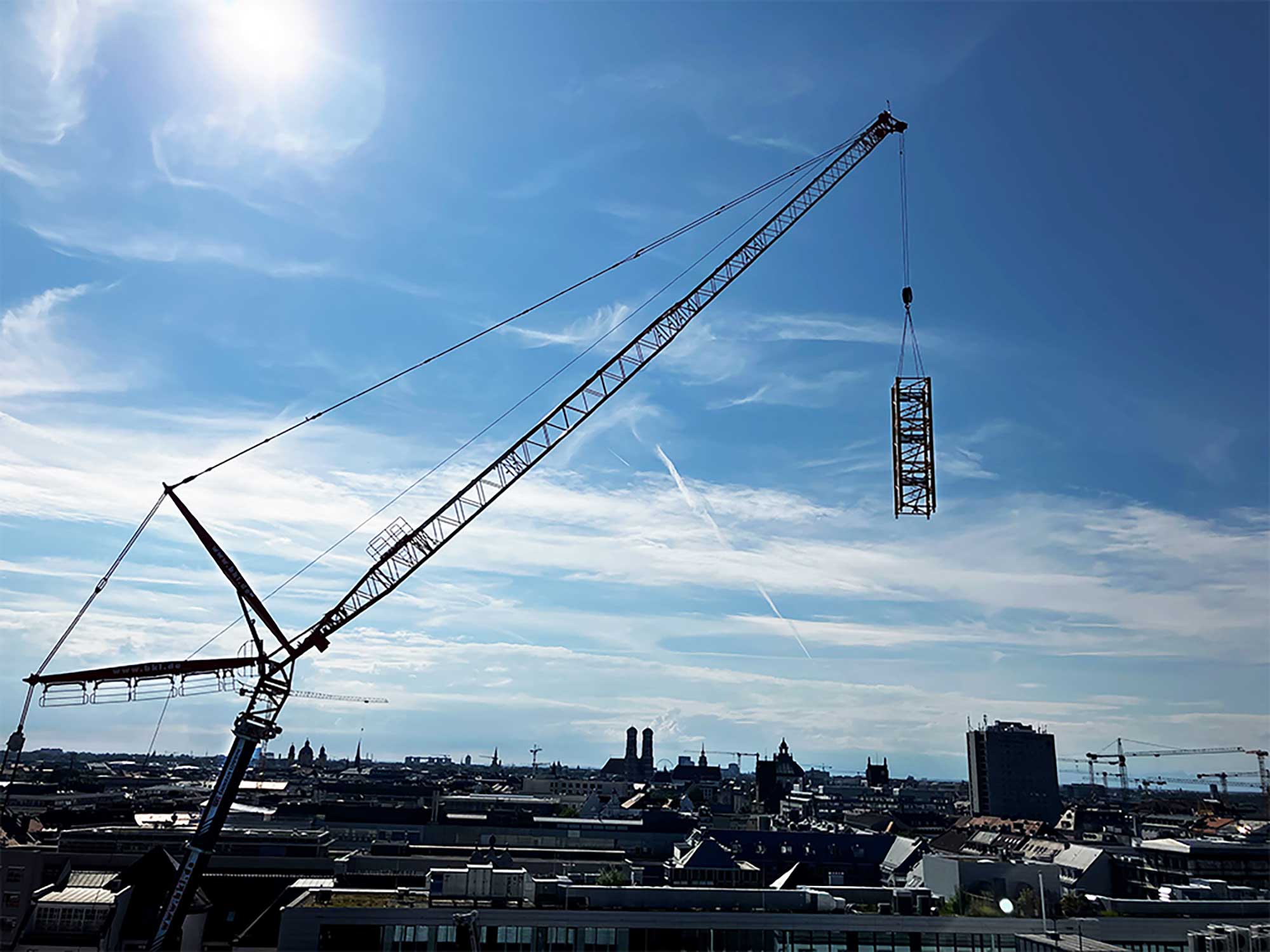 Baukran-Demontage in München: Liebherr Autokran LTM 1450-8.1 von BKL mit Wippspitze hebt Turmdrehkran TLS 80 20T Segment über Skyline, Demontage in der Luft bei Platzmangel in der Innenstadt, veranschaulicht Spezialisierung von BKL als Kran-Experten.