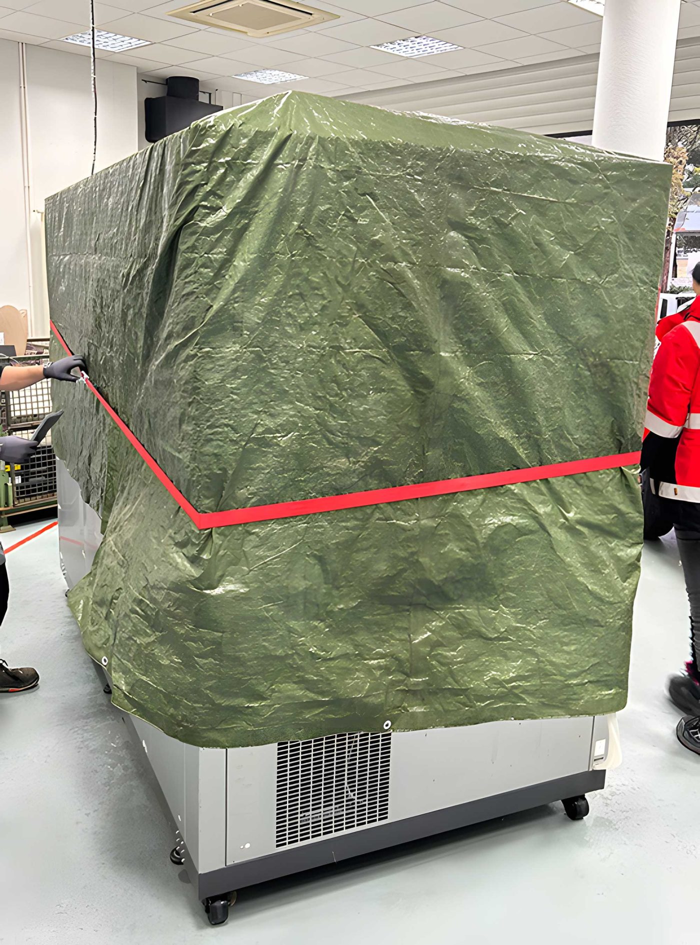 Eine große Industriemaschine, eingepackt in eine grüne Schutzplane und mit rotem Spanngurt gesichert, steht auf Rollen bereit für den Transport und Hub einer Industrieanlage durchgeführt von BKL Industriemontage