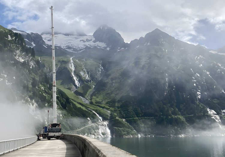 Ein Autokran LTM 1070-4.2 von BKL Rosenheim mit voll ausgefahrenem Teleskopausleger auf der schmalen Krone einer Staumauer am Stausee Zillergrund, eingebettet in eine beeindruckende Alpenlandschaft mit Wasserfällen und schneebedeckten Bergen, demonstriert präzisen Kran-Einsatz in beengten und anspruchsvollen Umgebungen für den BKL Blog.