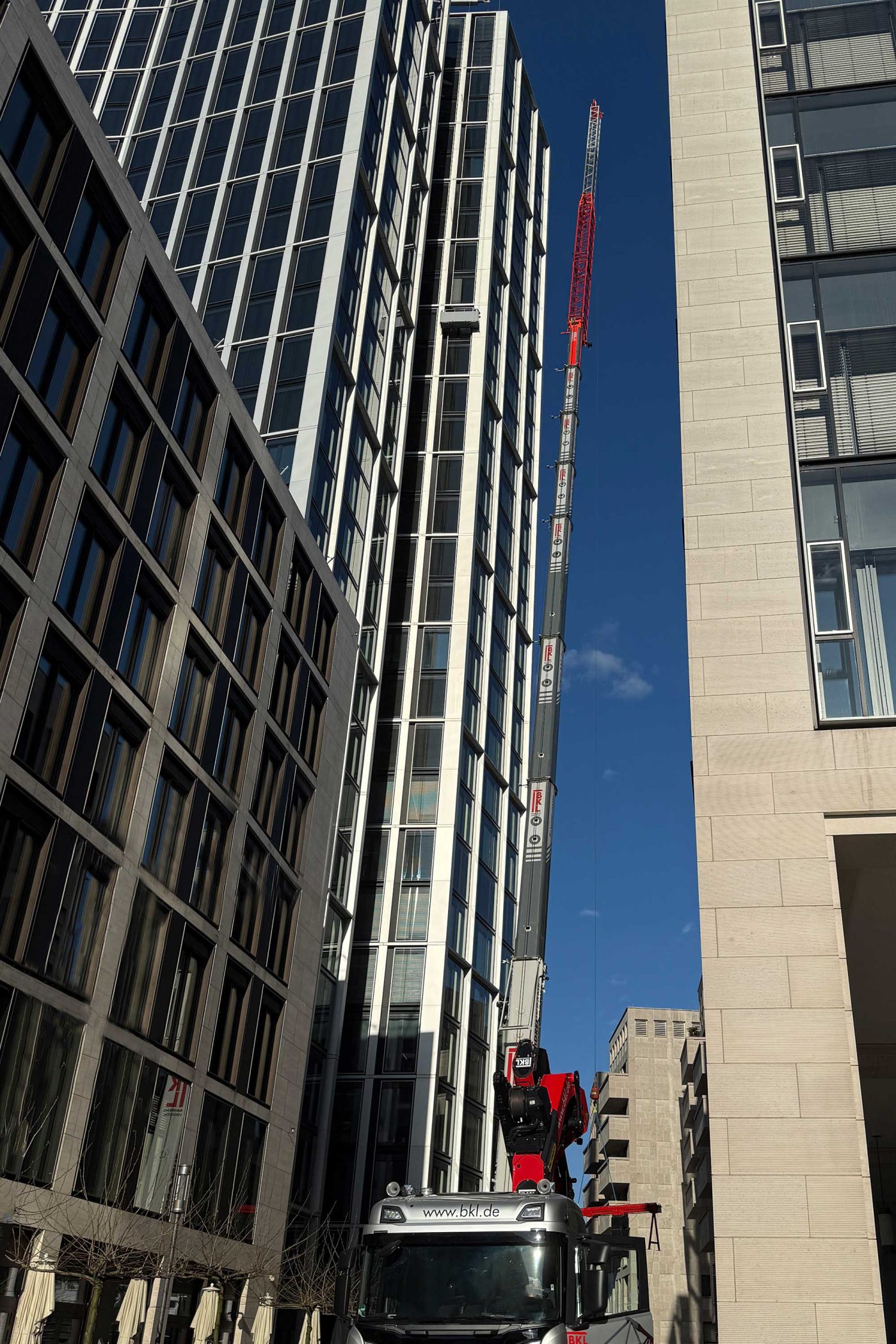 Ein 230-Tonnen Autokran LTM 1230-5.1 von BKL Frankfurt mit voll ausgefahrenem Teleskopausleger und roter Gitterverlängerung vor dem modernen WINX Tower Hochhaus, demonstriert Hochhaus-Kranarbeiten und Scheibentausch in extremer Höhe in der Frankfurter Innenstadt.