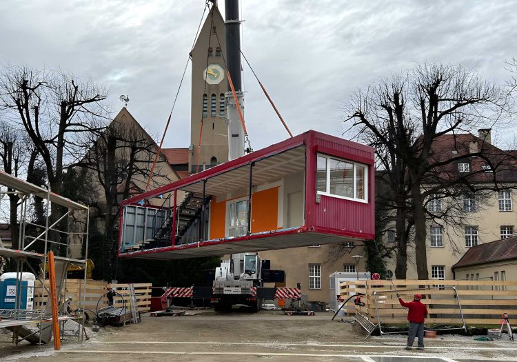 Ein rotes, langes Containermodul wird von einem Autokran LTM 1150-5.3 von BKL über einer Baustelle vor einer historischen Kirche positioniert, demonstriert knifflige Container-Montage und logistische Kran-Expertise für Modulbauten im BKL Blog.