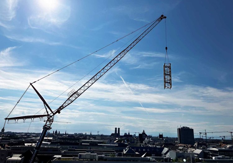 Ein großer Autokran von BKL Süd zerlegt den Ausleger eines Obendrehers in der Luft über der Münchner Skyline mit den Türmen der Frauenkirche, zeigt die komplexe Montage und Demontage von Turmdrehkranen und spezialisierte Kran-Einsätze bei beengten Platzverhältnissen.