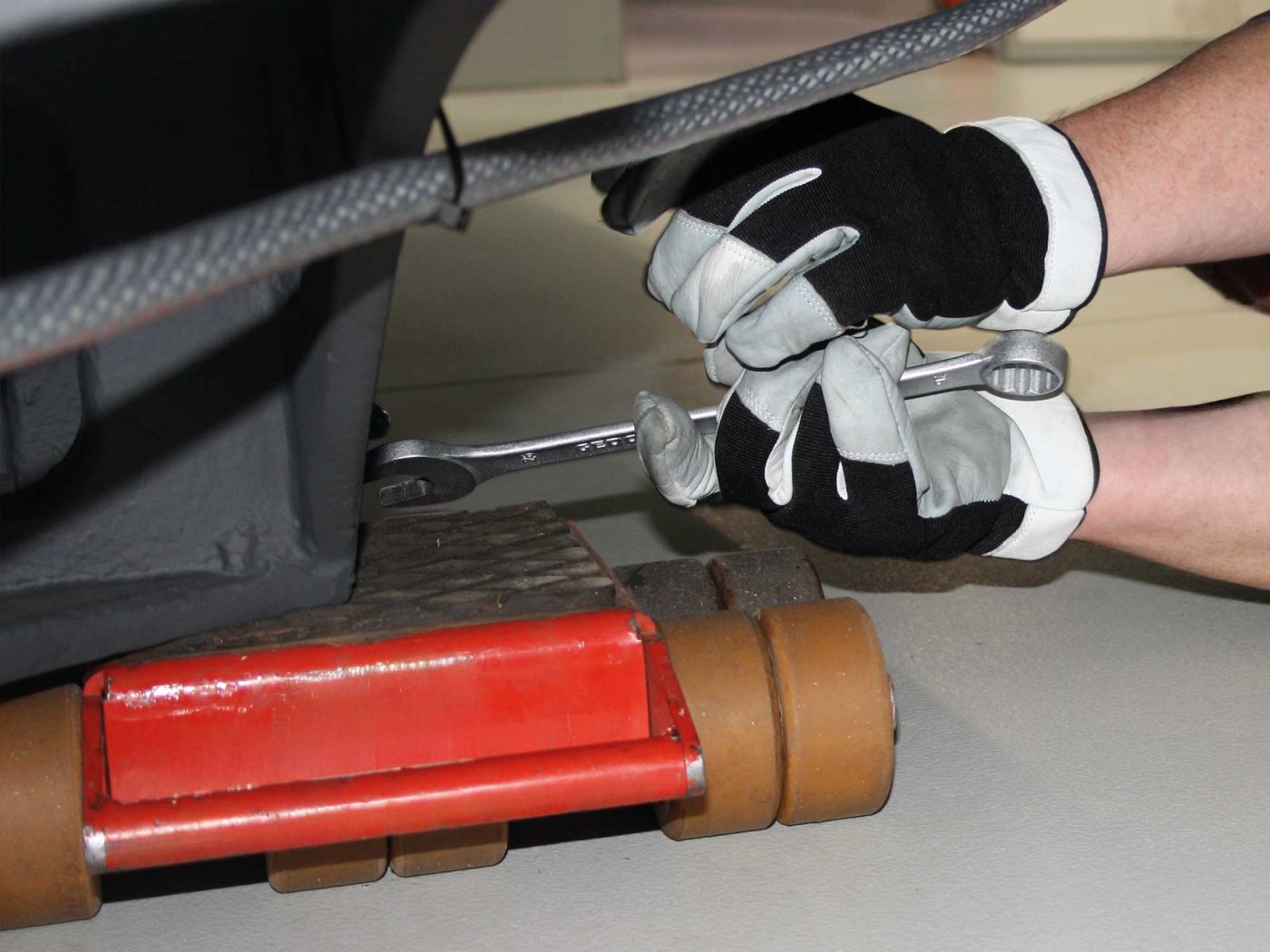 Detailaufnahme einer Industriemontage, bei der ein Mitarbeiter mit Schutzhandschuhen eine Schraube an einer schweren Maschine festzieht, die für den Maschinentransport auf Rollen positioniert ist.