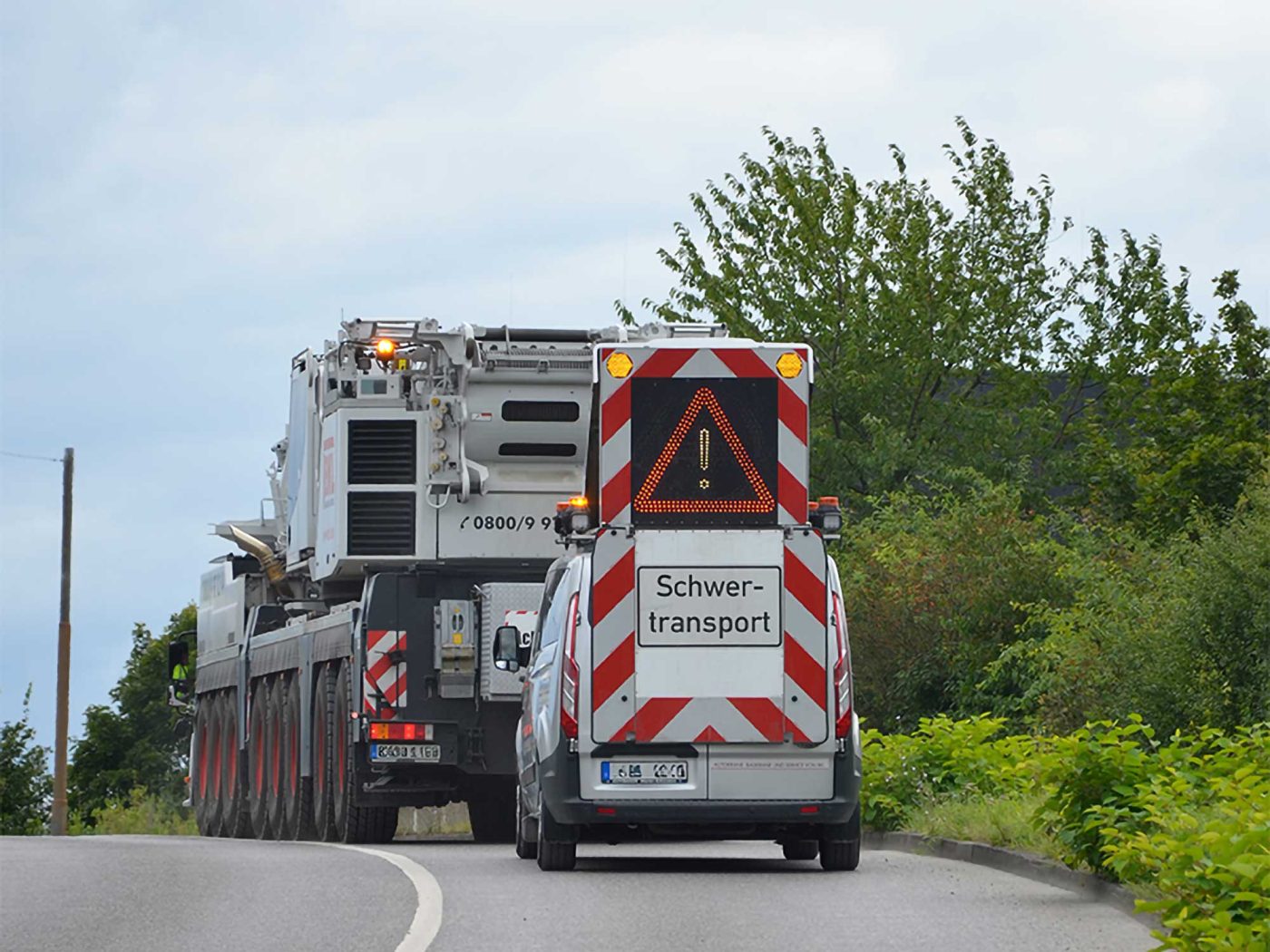 Ein schwerer BKL Krantransport mit mehreren Achsen, begleitet von einem BF-Begleitfahrzeug, das ein großes Warnschild mit Blinklicht und der Aufschrift "Schwertransport" zeigt, auf einer kurvigen Straße. Das Bild illustriert professionelle Transportbegleitung, Organisation von Streckenplanung, Genehmigungsmanagement, Sicherung und Logistik für Großraum- und Spezialtransporte.