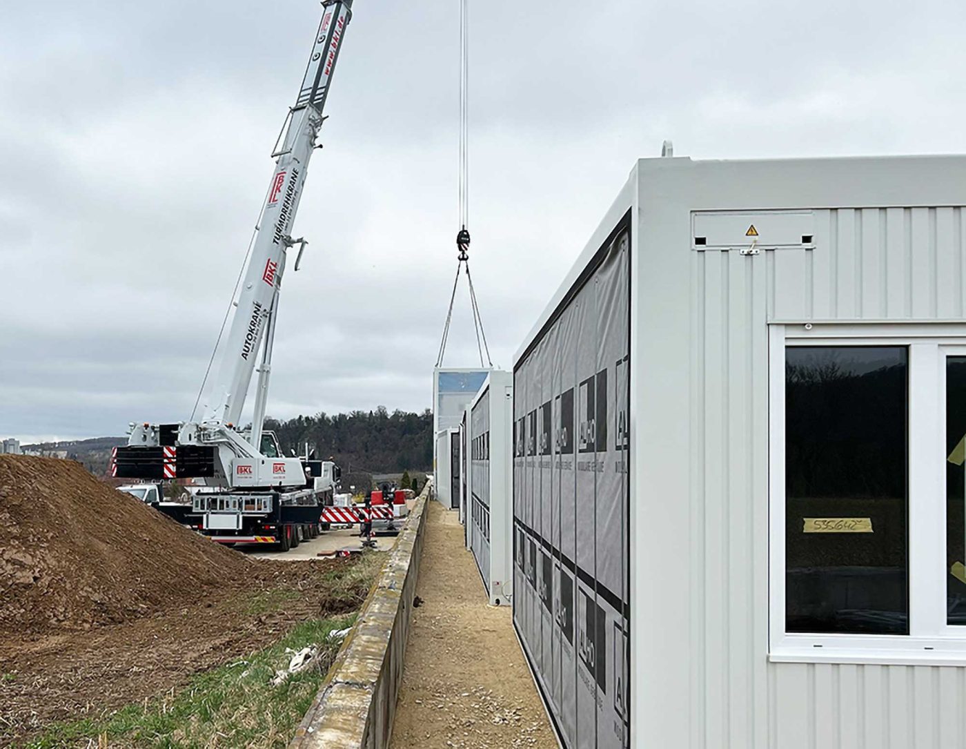 BKL Autokran bei der professionellen Containermontage: Ein weißer Mobilkran hebt einen Bürocontainer für den Bau einer modularen Containeranlage auf einer Baustelle, mit bereits installierten Modulen im Hintergrund.