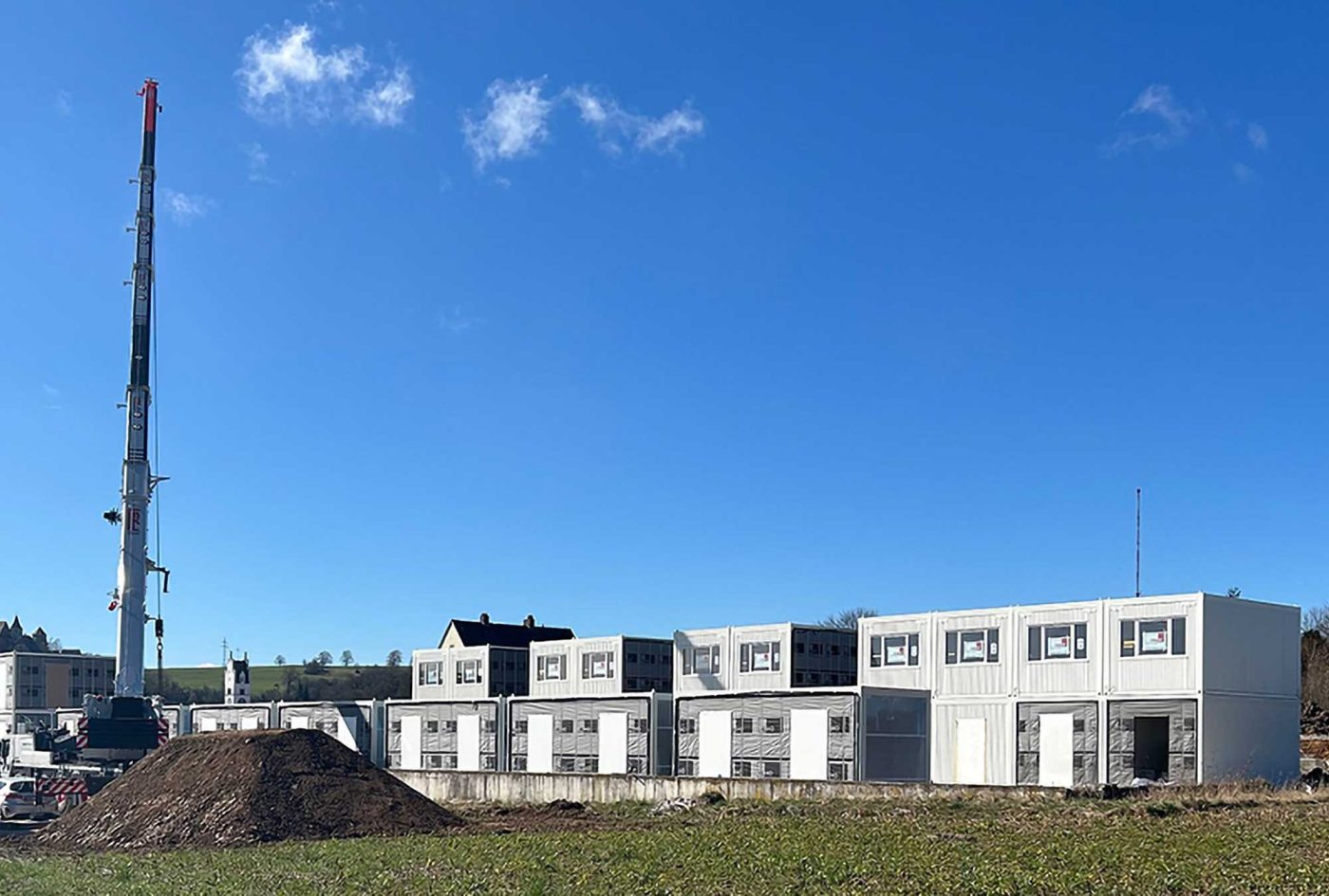 BKL Containermontage einer großen modularen Containeranlage, die mit einem Kran auf einer Baustelle unter strahlend blauem Himmel errichtet wird, geeignet als Bürocontainer, Wohncontainer oder Schulcontainer
