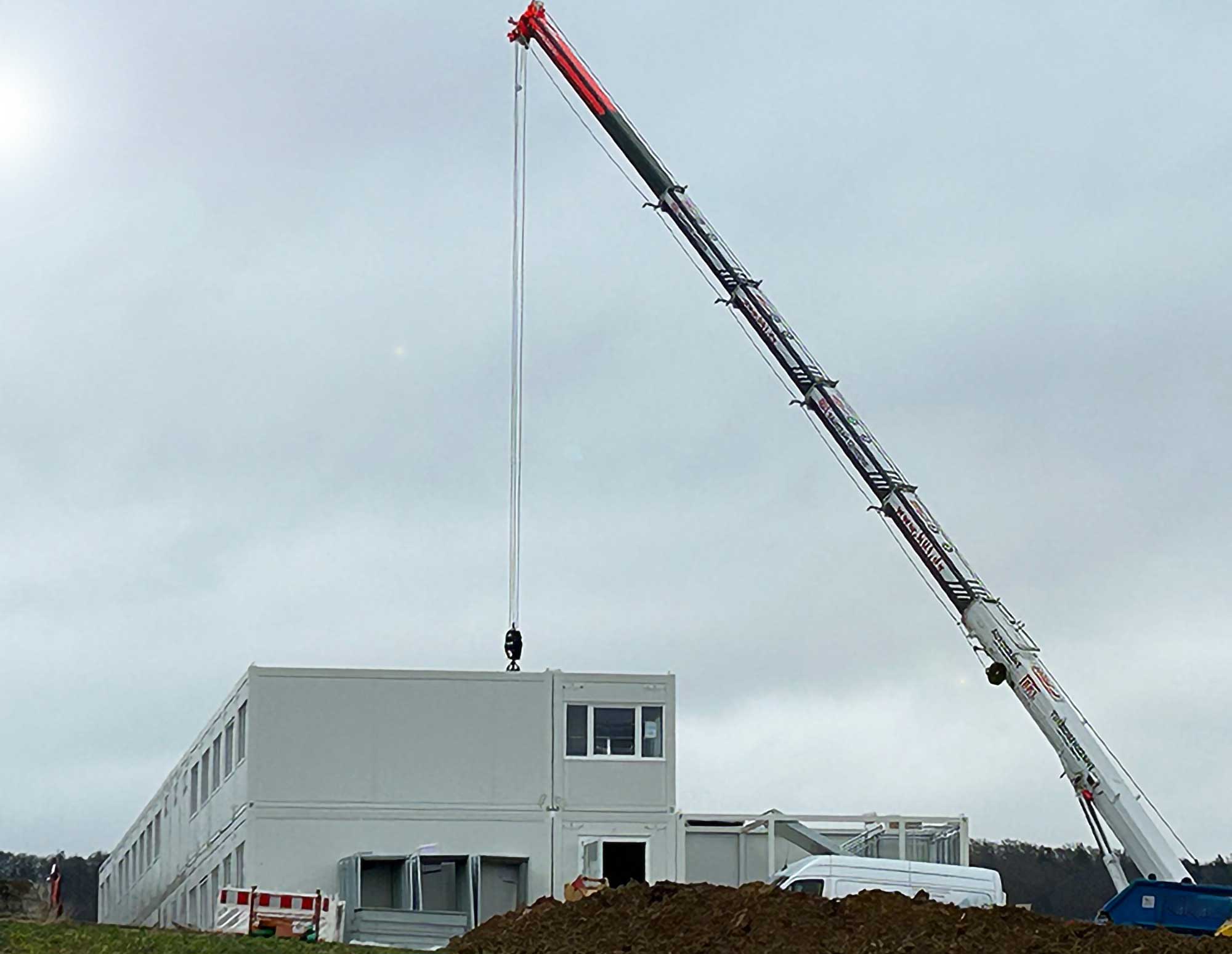 Ein Autokran montiert ein Raummodul für eine BKL Containeranlage im Modulbauweise, mit weiteren Containern im Hintergrund auf der Baustelle.