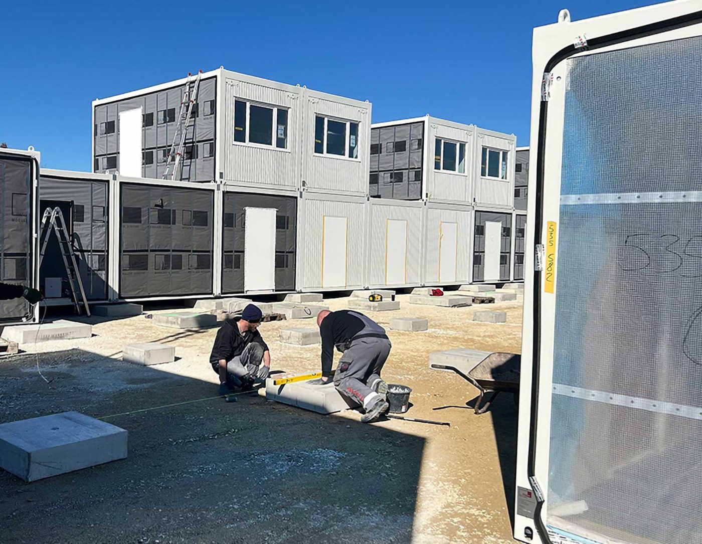 Zwei Bauarbeiter nivellieren Fundamente für die präzise Containermontage einer modularen Containeranlage, die aus gestapelten Büro- oder Wohncontainern besteht, unter blauem Himmel, stellvertretend für BKL Containermontage auf Baustellen.